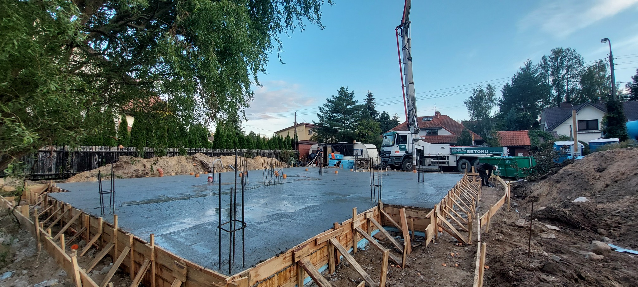 Wylany beton na fundamentach domu jednorodzinnego, widoczne szalunki, wystające pręty zbrojeniowe i pompa do betonu w tle.