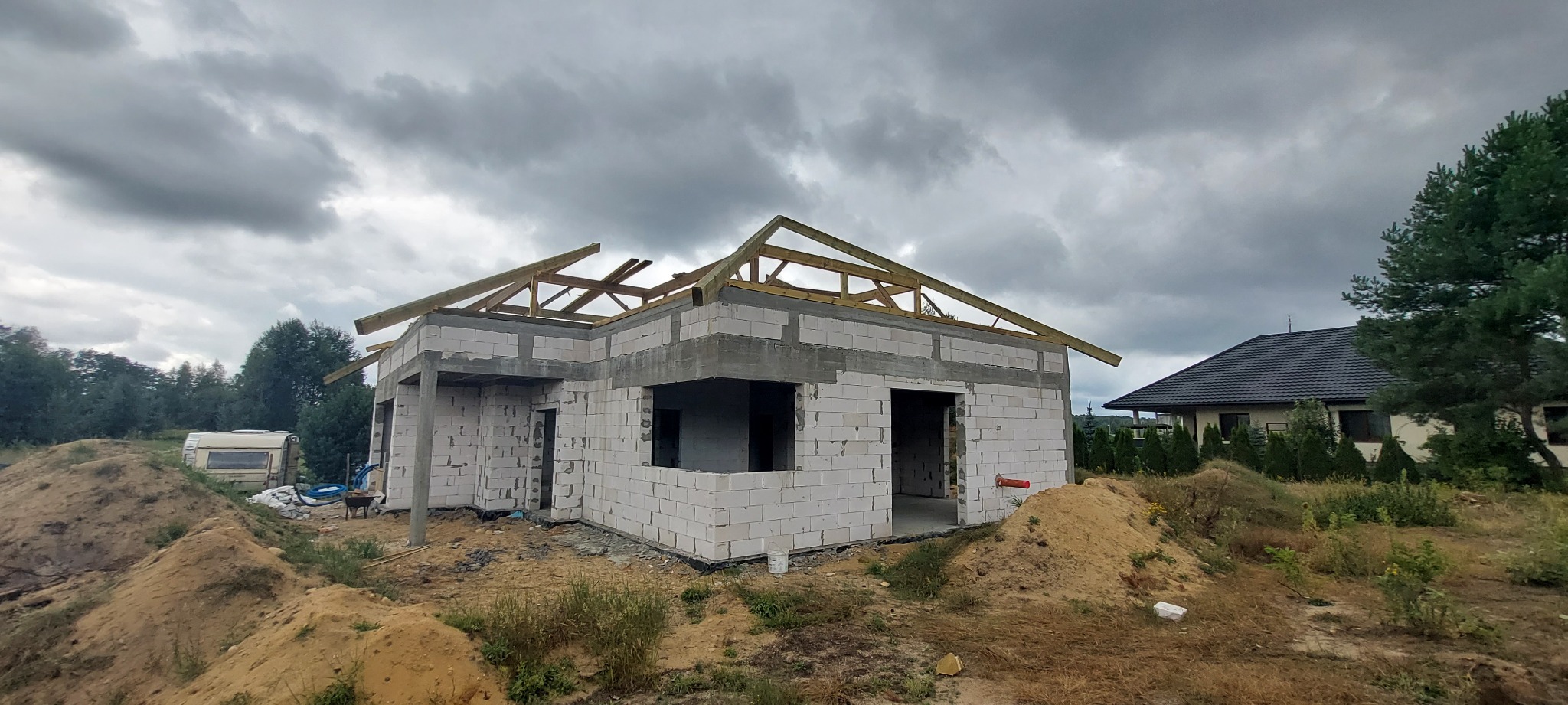 Surowy stan domu z białych bloczków, z drewnianą konstrukcją dachu w trakcie budowy, widok na sąsiedni dom i przyczepę kempingową w tle, pochmurne niebo.