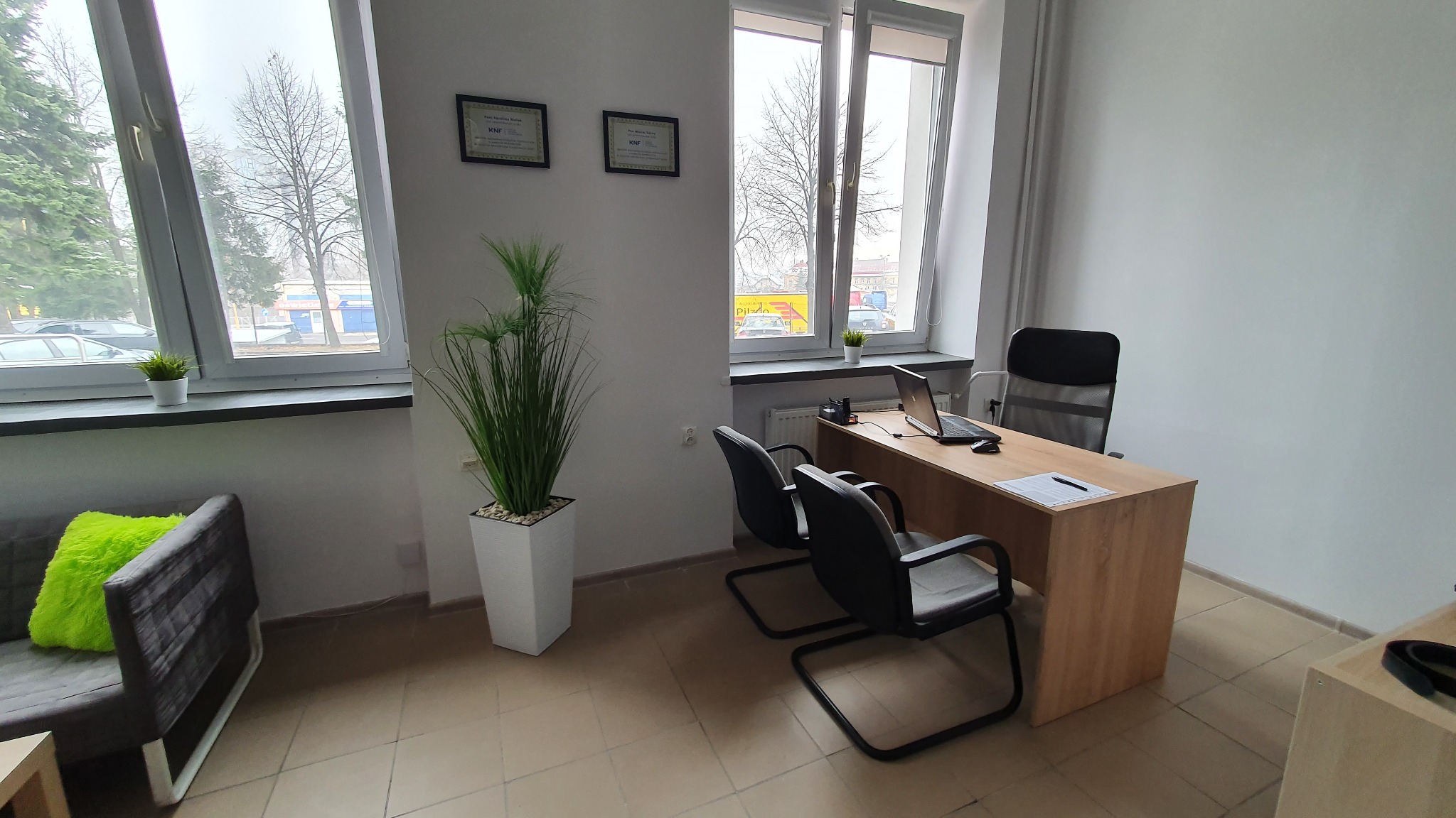 Biuro doradcy: biurko z laptopem, krzesła dla klientów, roślina doniczkowa, certyfikaty KNF na ścianie, widok z okna na ulicę.