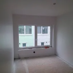 Wnętrze pokoju w trakcie wykańczania, ściany i sufit świeżo wyszpachlowane na gładko, widoczne narzędzia na parapecie okna, podłoga przygotowana pod dalsze prace.