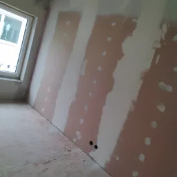 Wnętrze pokoju w trakcie prac wykończeniowych, widoczne ściany z nałożonymi pasami gładzi szpachlowej oraz widoczne otwory na gniazdka elektryczne.