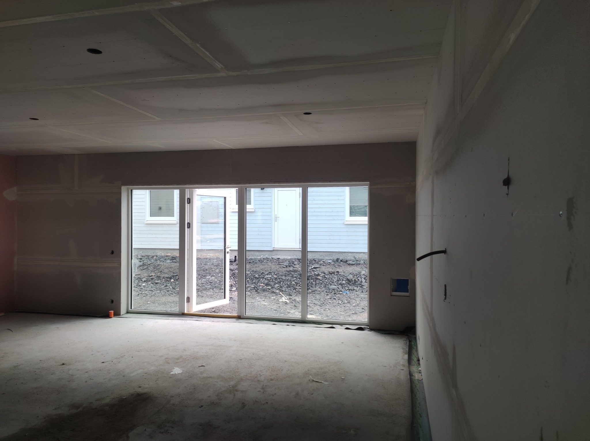 Surowe wnętrze z zamontowanymi płytami kartonowo-gipsowymi na ścianach i suficie, widoczne otwarte drzwi balkonowe z widokiem na podwórko z kamieniami.