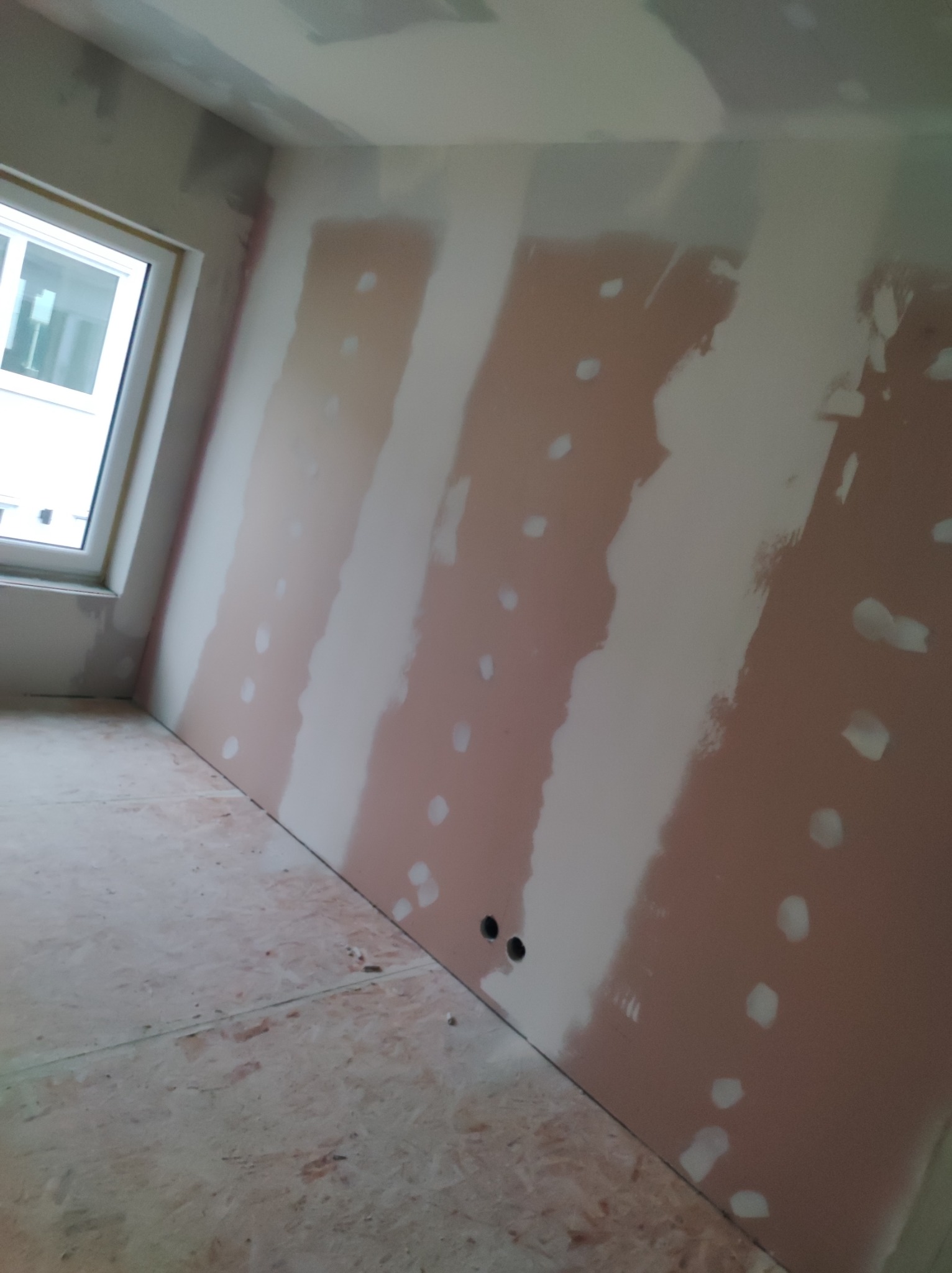 Wnętrze pokoju w trakcie prac wykończeniowych, widoczne ściany z nałożonymi pasami gładzi szpachlowej oraz widoczne otwory na gniazdka elektryczne.