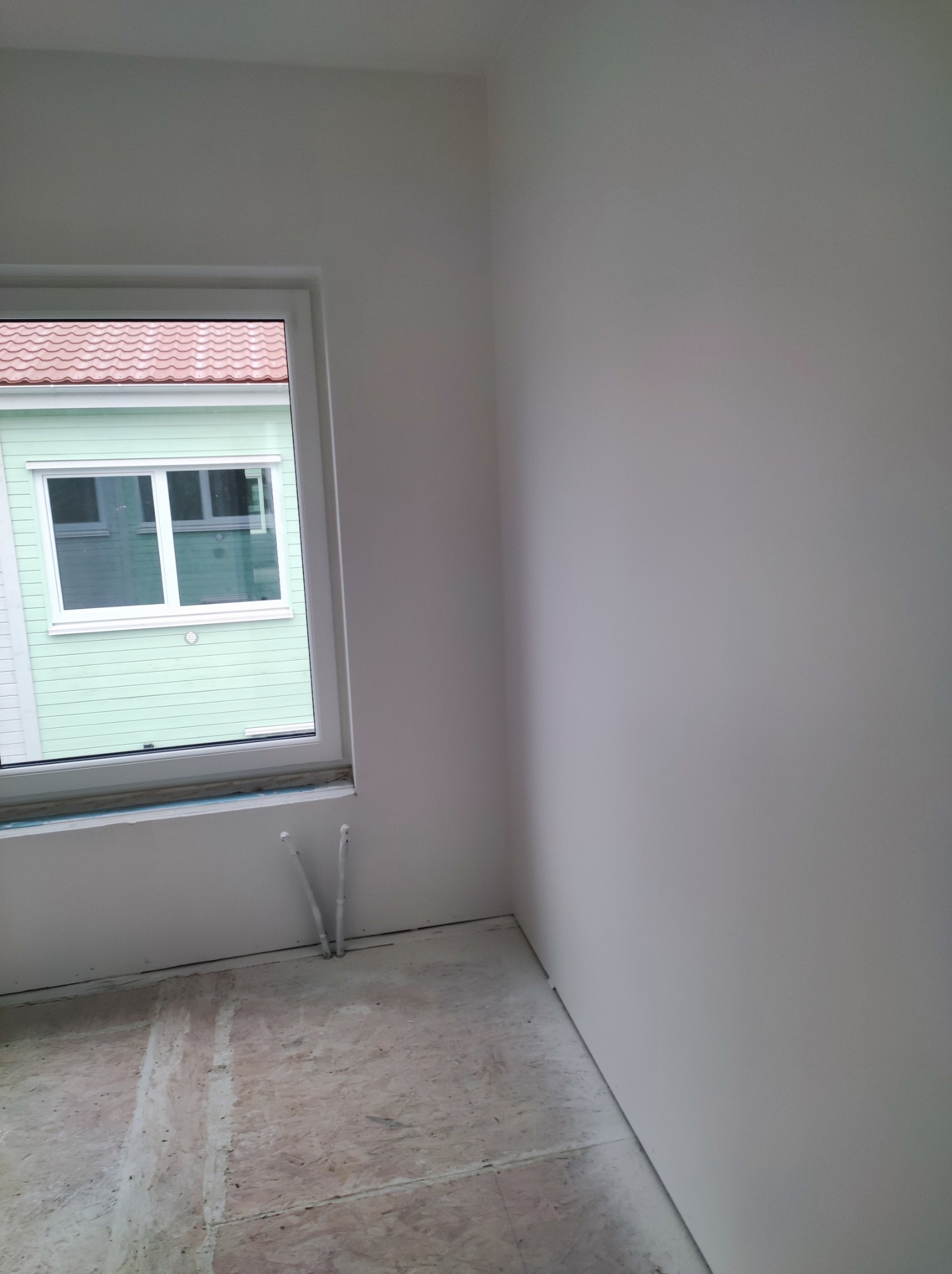 Pusty, pomalowany na biało pokój z widokiem na zielony dom z czerwonym dachem przez okno; widoczne ślady po szpachlowaniu na podłodze.