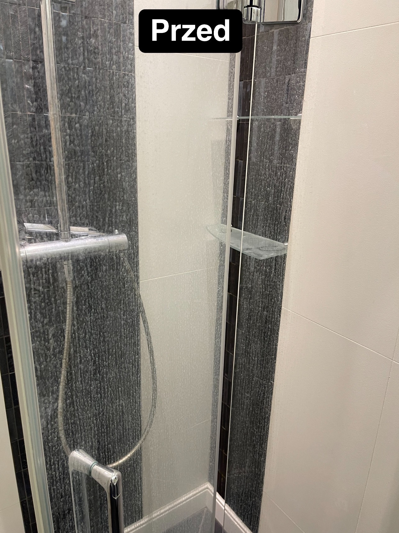 Zakamieniona kabina prysznicowa z widocznym osadem na szklanych drzwiach, czarnych płytkach i armaturze, z naklejonym napisem 'Przed' w górnym rogu.