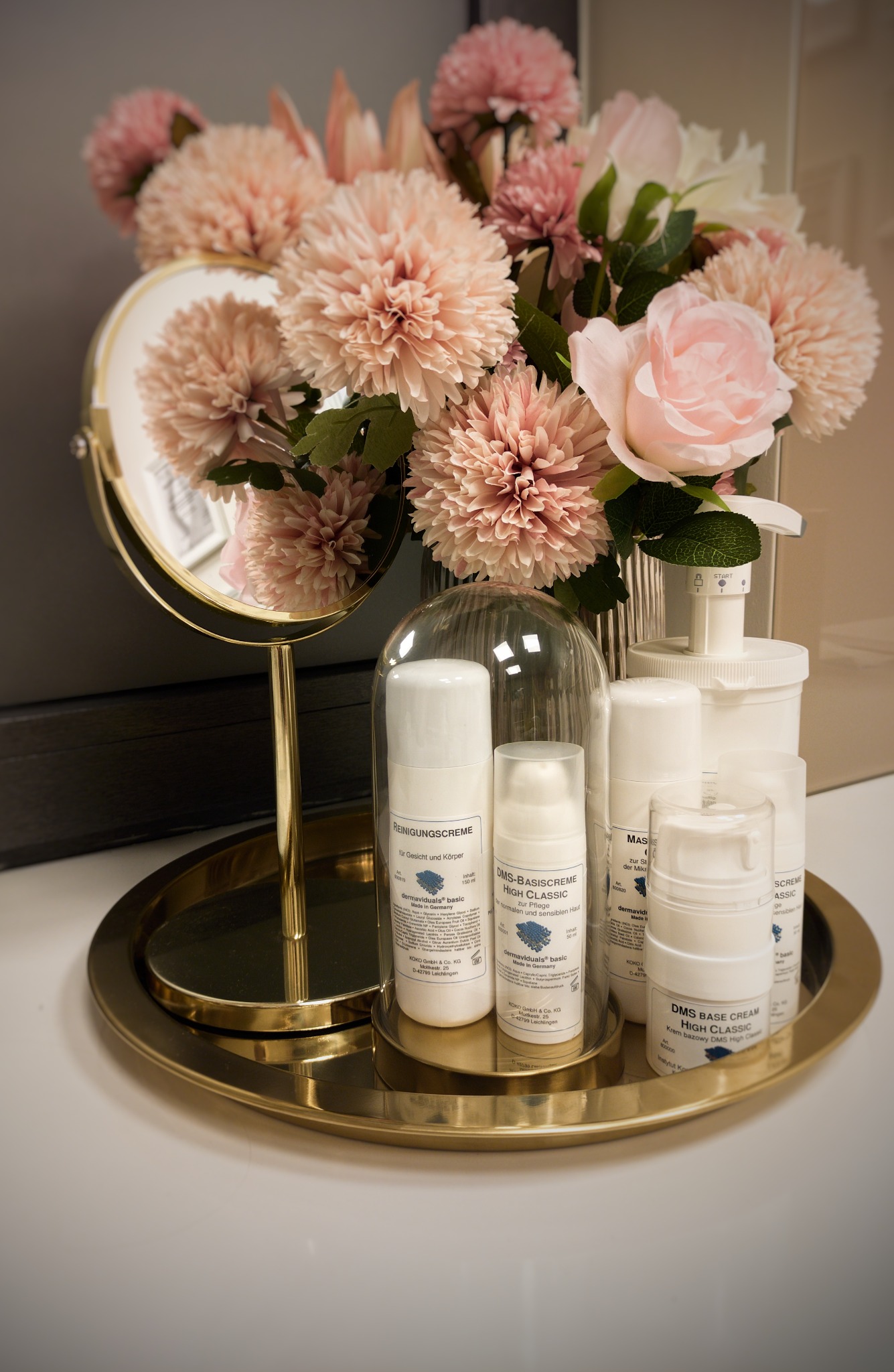 Zestaw kosmetyków marki dermaviduals na złotej tacy obok lustra i bukietu kwiatów w odcieniach różu.