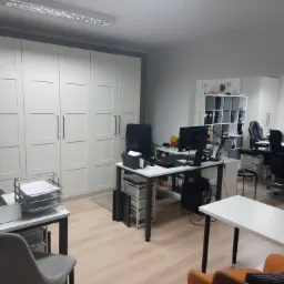 Jasne biuro z białymi meblami, biurkami z komputerami i krzesłami biurowymi, widok na przestrzeń do pracy.