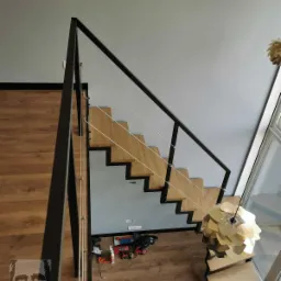 Widok z góry na nowoczesne schody z drewnianymi stopniami i czarną metalową balustradą z linkami, na podłodze narzędzia i logo firmy.