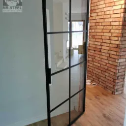 Szklane drzwi z czarną metalową ramą, podzielone na kwadraty, otwierają się do wnętrza z widocznymi schodami i ceglanym filarem, drewniana podłoga, logo firmy widoczne na ścianie.