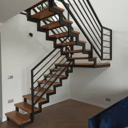 Nowoczesne schody o konstrukcji metalowej z drewnianymi stopniami, czarne poręcze, widok z dołu, parkiet w jodełkę na podłodze, fragment niebieskiej sofy w rogu.