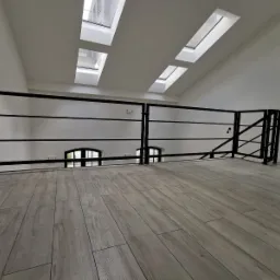 Metalowa balustrada antresoli z poziomymi prętami i schodami, widok z góry na drewnopodobną podłogę i okna dachowe w białym pomieszczeniu.