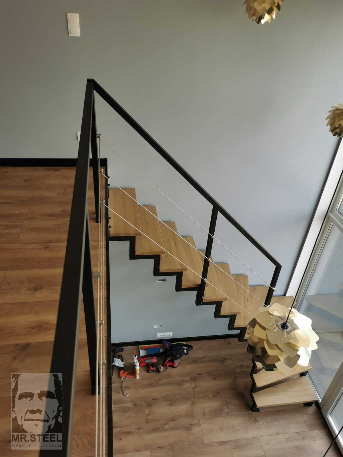 Widok z góry na nowoczesne schody z drewnianymi stopniami i czarną metalową balustradą z linkami, na podłodze narzędzia i logo firmy.