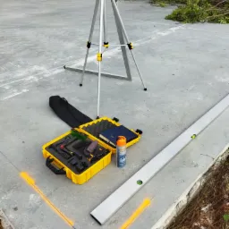 Laserowy niwelator na statywie, poziomica, żółta skrzynka z narzędziami i pomarańczowy spray do znakowania na surowym betonowym podłożu z wyznaczonymi liniami.