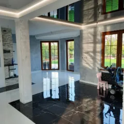 Wykończone wnętrze z czarno-białą podłogą, szare ściany z fakturą, podwieszany sufit z oświetleniem LED i widocznymi pęknięciami na suficie odbitymi w lustrzanej powierzchni.