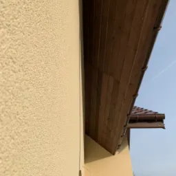 Fragment elewacji budynku z widocznym ociepleniem i podbitką dachową w odcieniach brązu, uchwycony pod kątem w pochmurny dzień.