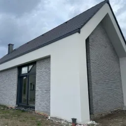 Nowoczesny dom jednorodzinny z szarej cegły klinkierowej i białym tynkiem, czarna stolarka okienna i dachówka, widok z zewnątrz w pochmurny dzień.