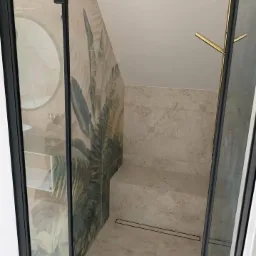 Nowoczesna kabina prysznicowa z czarnymi profilami, złotą armaturą i płytkami z motywem tropikalnych liści na ścianie.