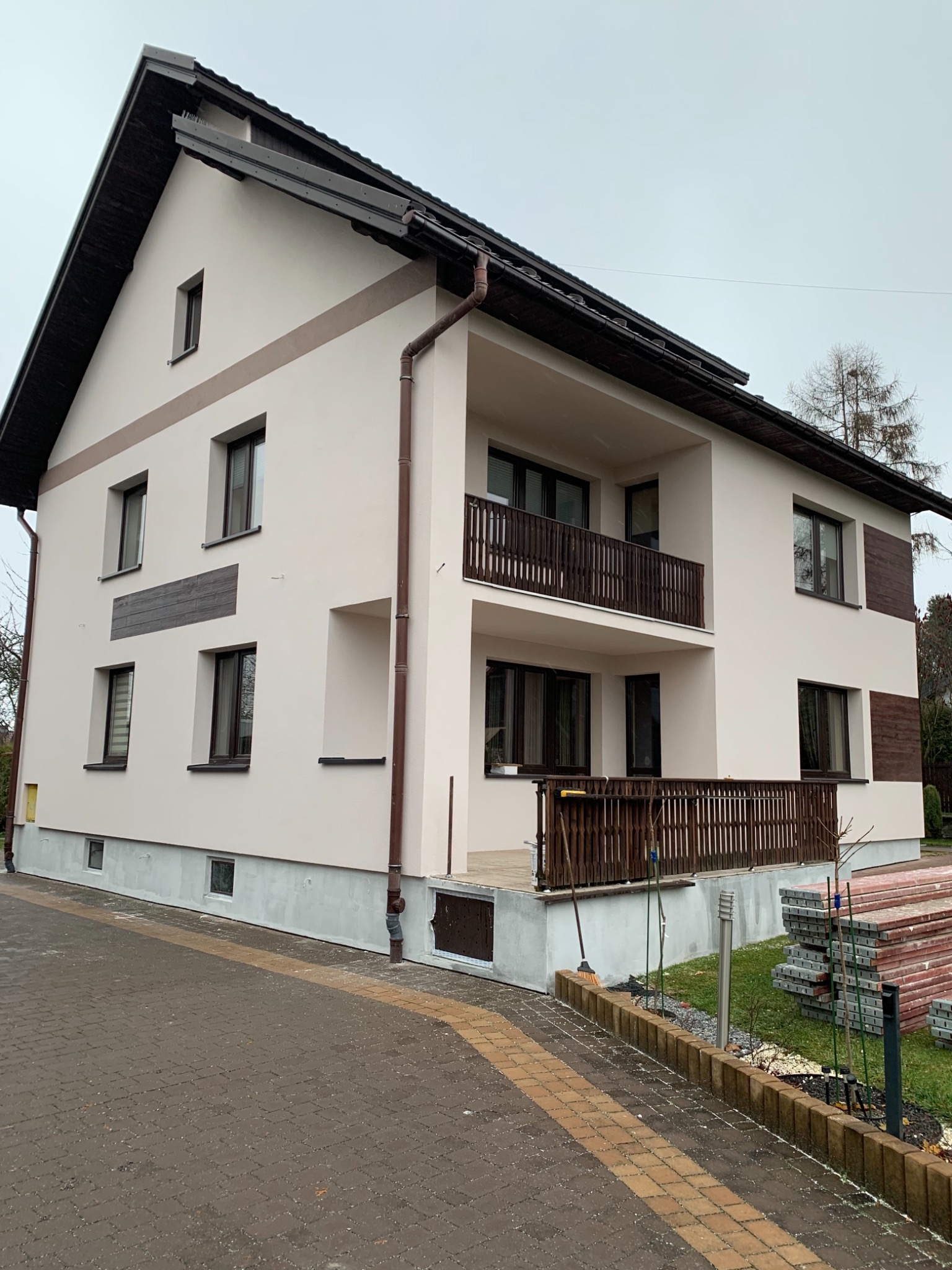 Dwupiętrowy dom z balkonami, świeżo po remoncie elewacji, z widocznymi elementami dekoracyjnymi z drewna i stertą materiałów budowlanych obok