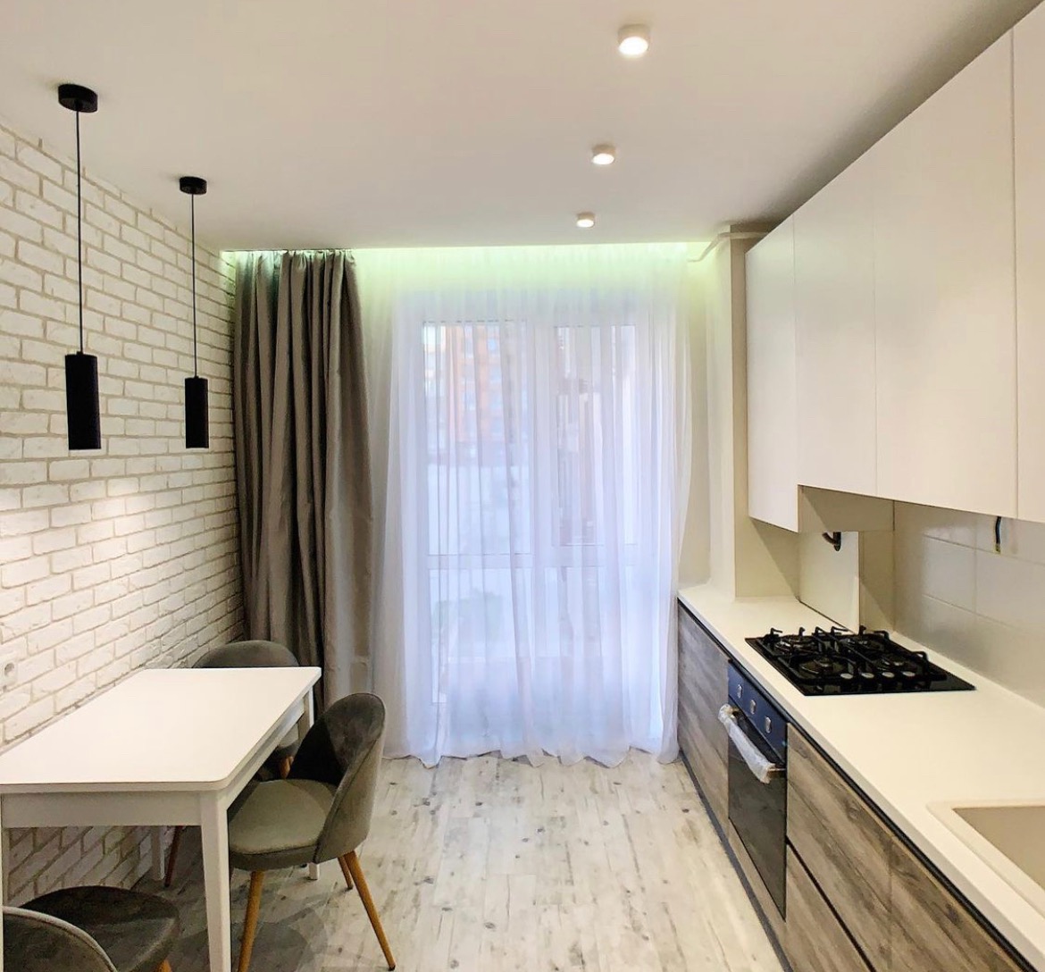 Jasna kuchnia z białymi szafkami, drewnopodobnymi frontami, białym stołem i szarymi krzesłami w stylu skandynawskim, oświetlona lampami wiszącymi i punktowymi, z widokiem na okno zasłonięte firanką.