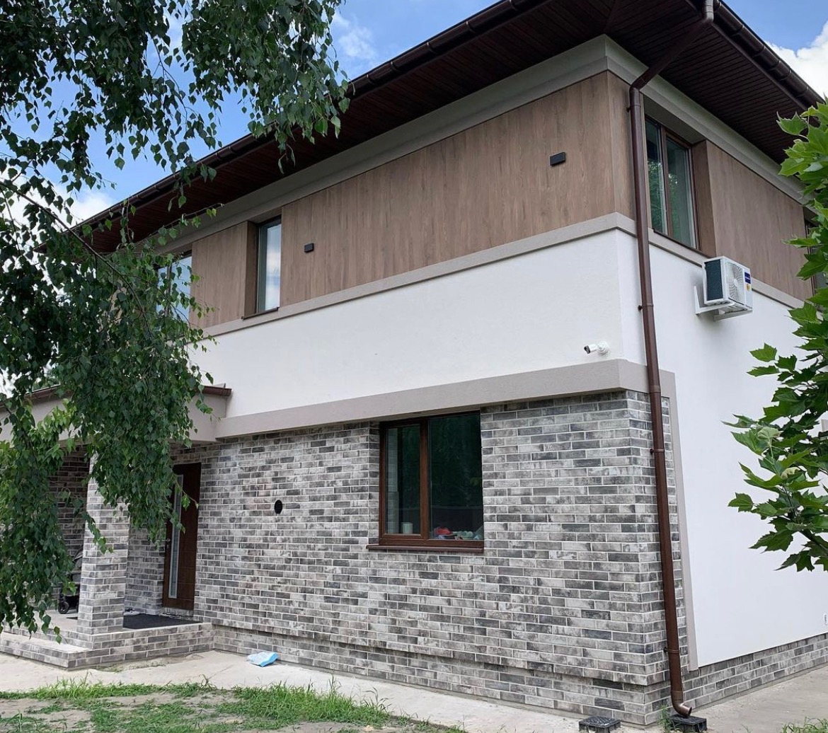 Dwukondygnacyjny dom z szarą cegłą klinkierową na parterze, białą elewacją na piętrze i drewnianą okładziną pod dachem, widoczny klimatyzator na ścianie, rynna i zieleń w tle.