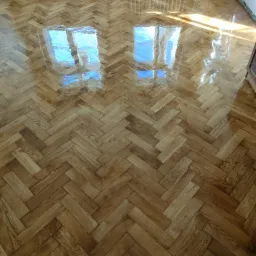 Parkiet ułożony w jodełkę, świeżo polakierowany, z odbiciem okna, w pokoju z grzejnikiem i fragmentami ściany w stanie surowym.