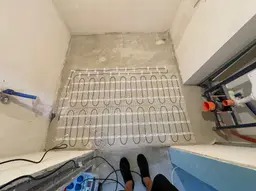 Rozłożona mata grzewcza na podłodze w łazience przed wylewką, widoczne przewody i elementy instalacji hydraulicznej na ścianach.