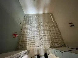 Rozłożona mata grzewcza na podłodze w trakcie montażu, widoczne kable i elementy instalacji elektrycznej, surowy stan wykończenia wnętrza.