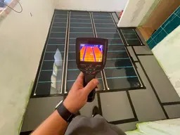 Sprawdzanie rozłożenia maty grzewczej termowizją przed zalaniem wylewką, widoczne przewody grzewcze pod folią i obraz termowizyjny na ekranie urządzenia.