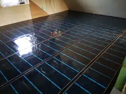 Rozłożona mata grzewcza z widocznymi niebieskimi przewodami na podłodze w trakcie instalacji. Kable elektryczne zwisają ze ściany w tle.
