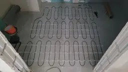 Rozłożona mata grzewcza na podłodze w trakcie montażu, widoczne kable, białe ściany w tle, częściowo położona wylewka.