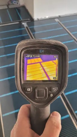 Pomiar temperatury termowizyjnej zainstalowanej podłogowej instalacji grzewczej, widoczne rury grzewcze pod podłogą na ekranie kamery termowizyjnej, w tle fragment pomieszczenia z białą ścianą.