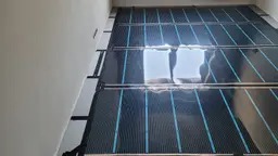 Rozłożona mata grzewcza z niebieskimi przewodami na podłodze, widok z góry pod kątem, surowy stan przed wylewką.