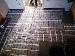 Rozłożona mata grzewcza na podłodze przed montażem, widoczne przewody grzejne i taśmy mocujące, fragment nogi instalatora w tle.