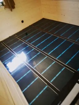 Rozłożona na podłodze czarna mata z niebieskimi elementami grzewczymi, widoczne białe ściany i czarny włącznik na ścianie.