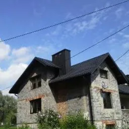 Dom jednorodzinny z nowym, ciemnym dachem z dachówki ceramicznej, widoczny komin, elewacja z kamienia, błękitne niebo z chmurami w tle.