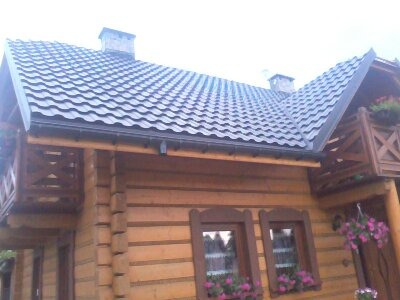 Dom z bali z nowym, ciemnoszarym dachem z blachodachówki, widoczne rynny i komin. Na balkonie i w oknach kwiaty.