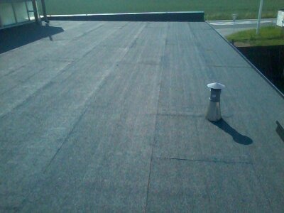 Płaski dach pokryty papą termozgrzewalną z kominkiem wentylacyjnym, widok z góry, na drugim planie budynek i zieleń.