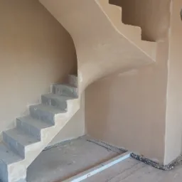 Wnętrze w trakcie prac: otynkowane ściany klatki schodowej ze schodami w surowym stanie. Widoczny poziomica na podłodze i kable przy ścianie.