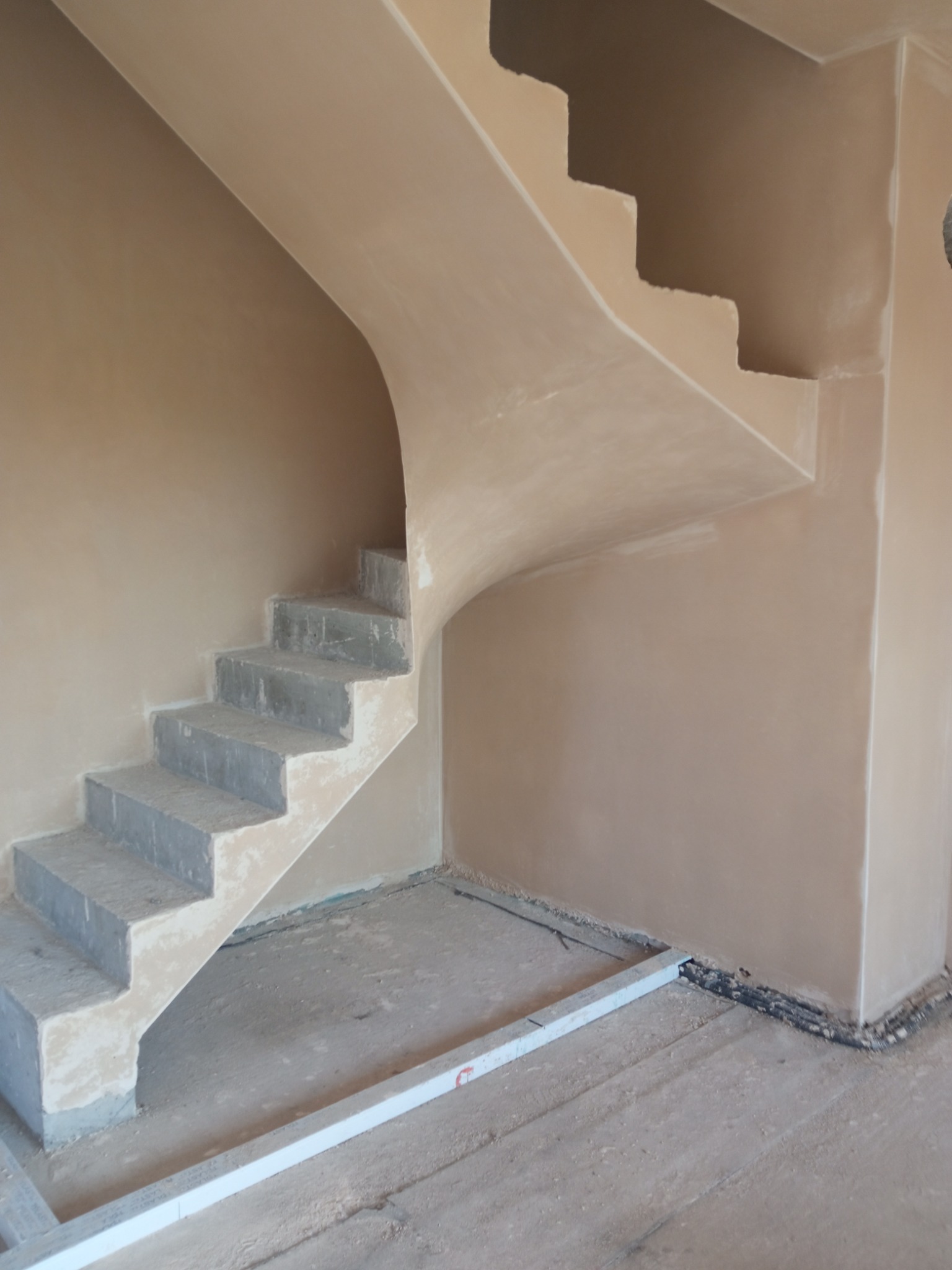 Wnętrze w trakcie prac: otynkowane ściany klatki schodowej ze schodami w surowym stanie. Widoczny poziomica na podłodze i kable przy ścianie.