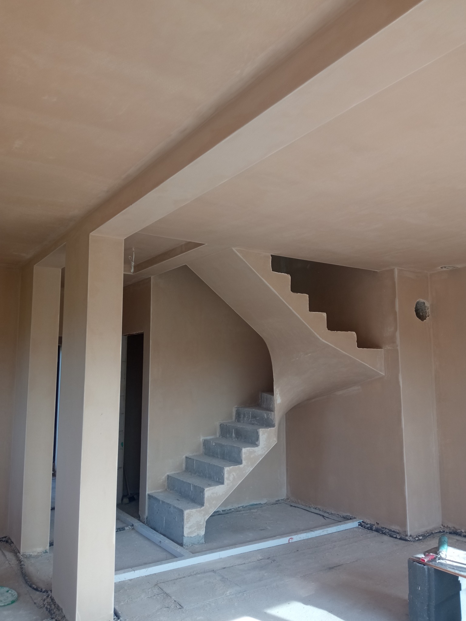 Wnętrze po tynkowaniu: świeżo otynkowane ściany, sufit i schody z widocznymi niedoskonałościami wykończenia. Widoczne filary i przygotowane podłoże pod podłogę.