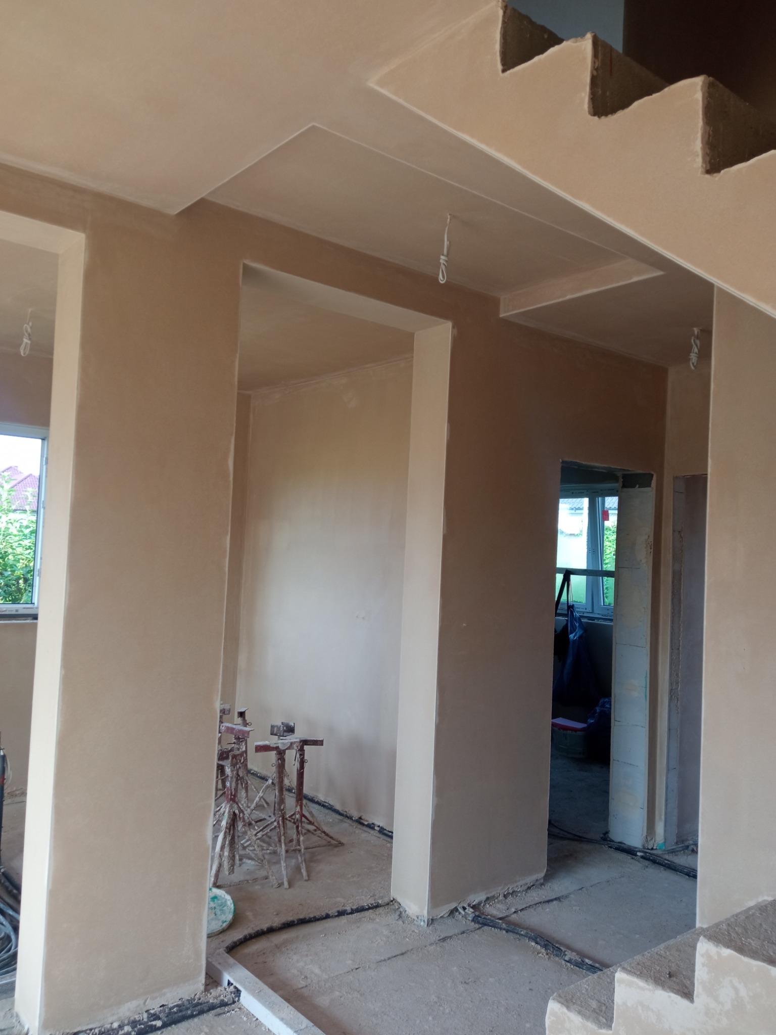 Wnętrze w trakcie prac wykończeniowych: świeżo otynkowane ściany, widoczne podpory, elementy schodów. Neutralna kolorystyka tynków.