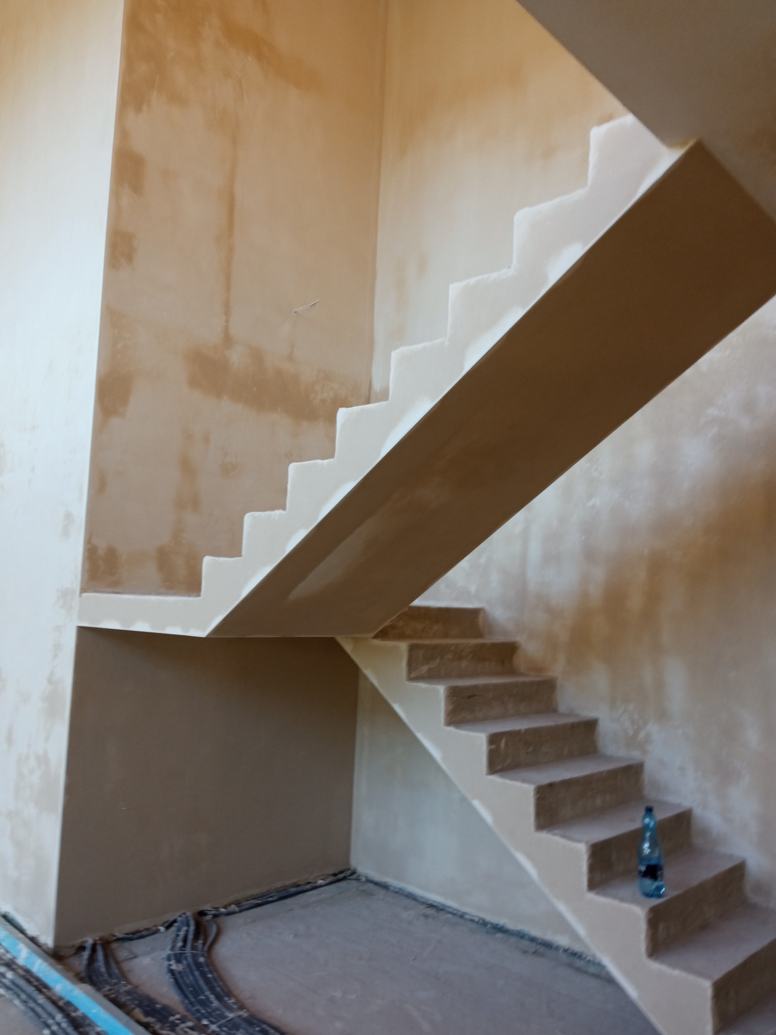 Wnętrze w trakcie prac tynkarskich: surowe ściany, fragment schodów z widoczną butelką wody. W tle widoczne kable instalacyjne.