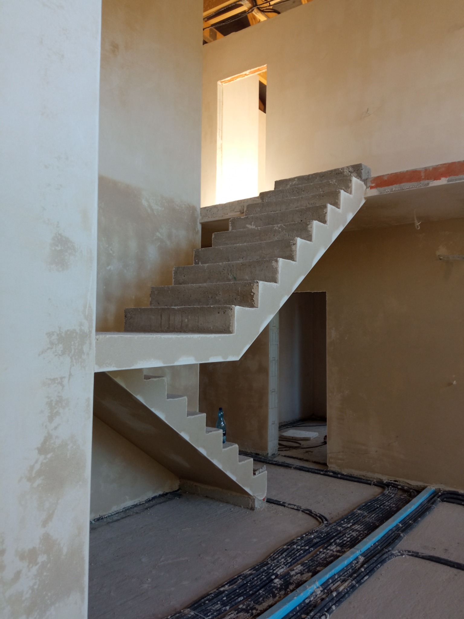 Surowe, betonowe schody w trakcie budowy, z widocznymi instalacjami na podłodze i świeżo nałożonymi tynkami na ścianach. W tle widoczne otwory drzwiowe.
