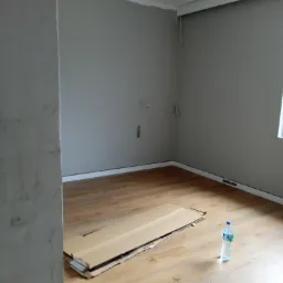 Pokój w Kaliszu w trakcie remontu: świeżo położona podłoga z paneli, kartony na podłodze, ściany przygotowane do malowania, widoczna butelka wody.