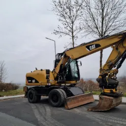 Żółta koparka kołowa Caterpillar M315D z łyżką i lemieszem stoi na asfalcie, widoczne ślady opon, w tle drzewa bez liści i pochmurne niebo.