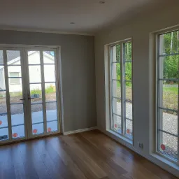 Jasny, przestronny pokój z drewnianą podłogą, białymi drzwiami balkonowymi i oknami z widokiem na ogród z drzewami i żwirową ścieżką. Ściany pomalowane na szaro i kremowo.