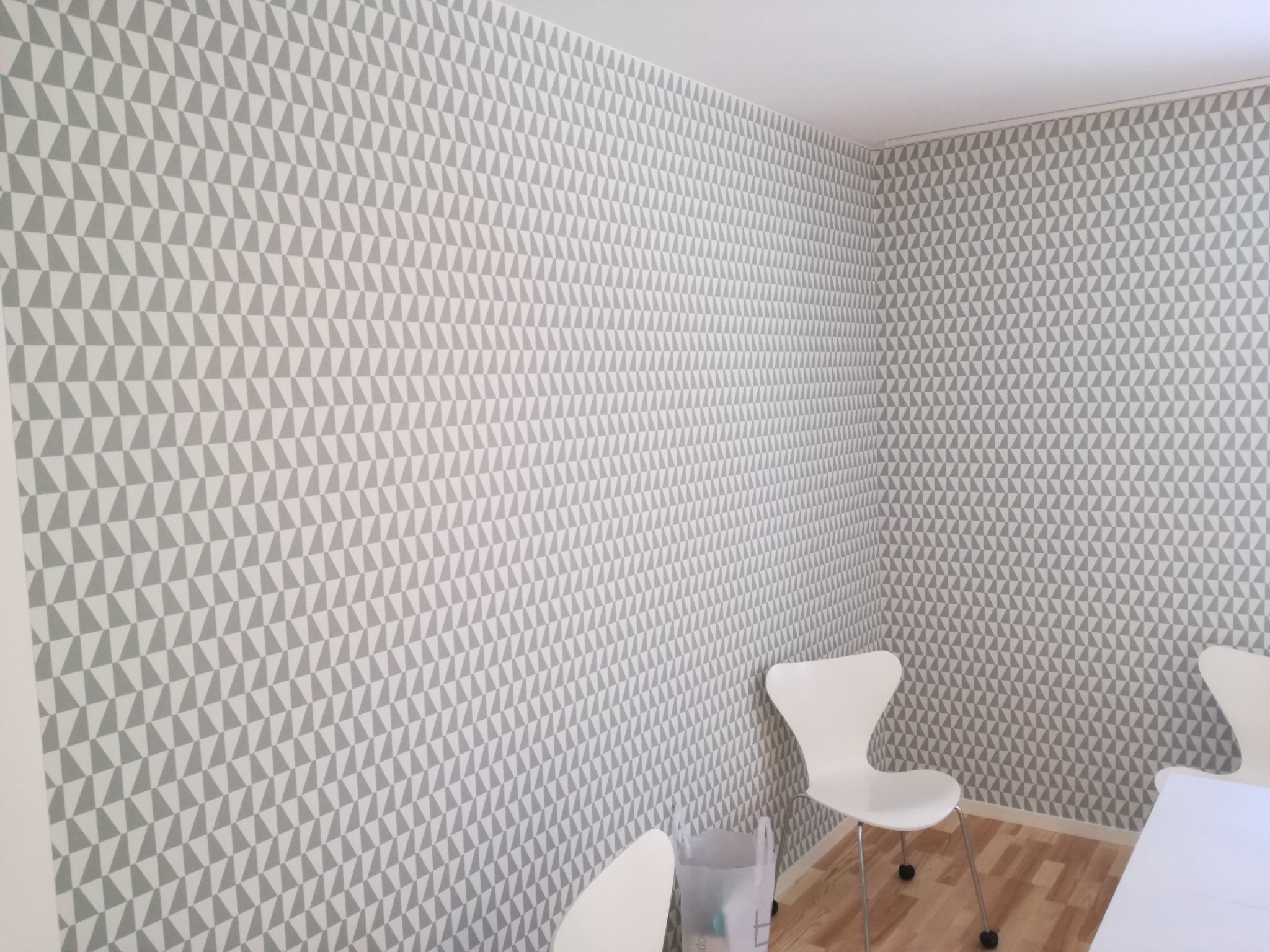 Ściana pokryta tapetą w szaro-białe trójkąty, widoczne białe krzesła i fragment blatu stołu w rogu pokoju.