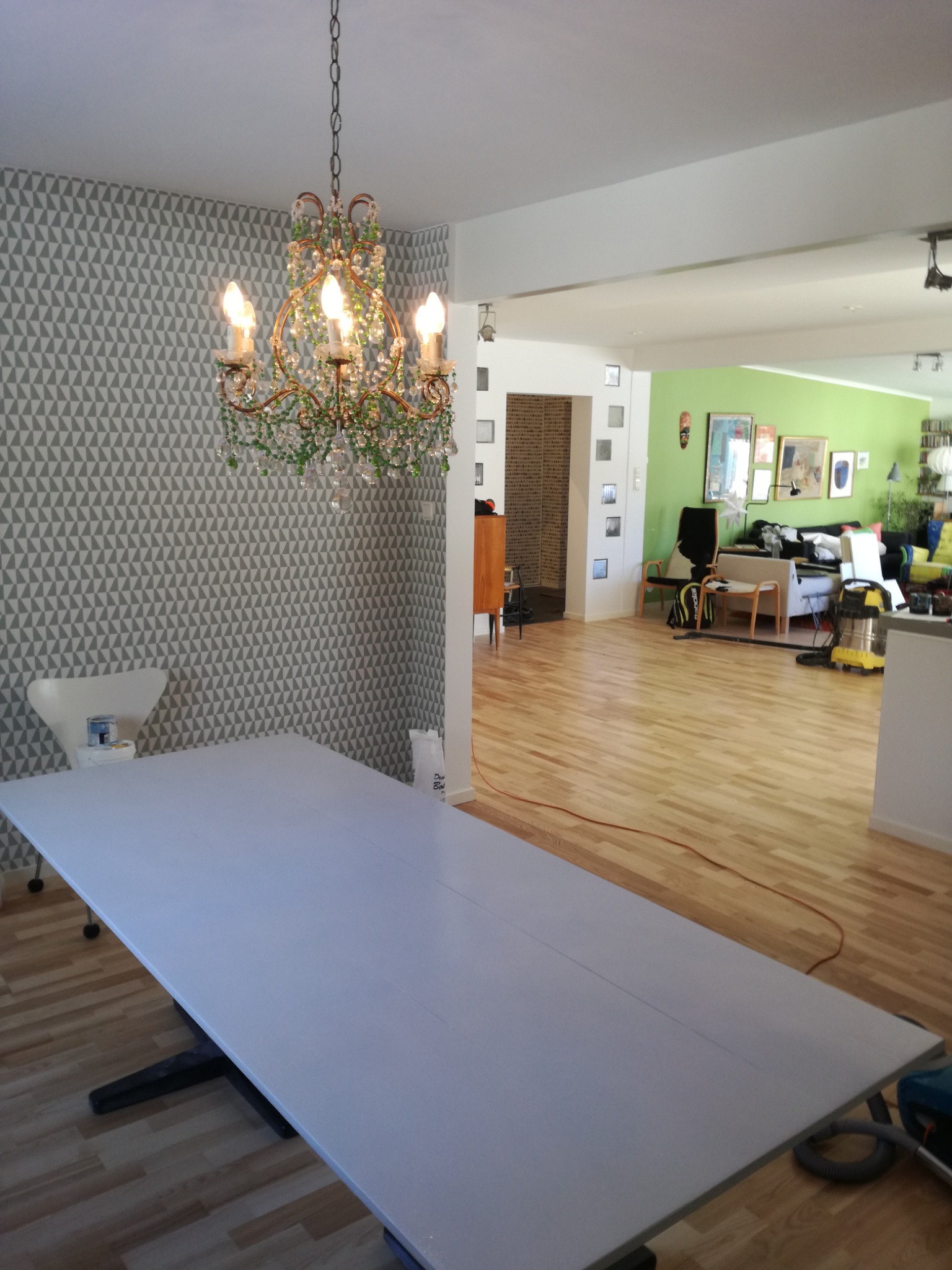 Pokój z odświeżoną tapetą w geometryczne wzory, centralnie umieszczony duży stół przygotowany do malowania, nad nim ozdobny żyrandol, w tle widoczny inny pokój z parkietem i meblami.