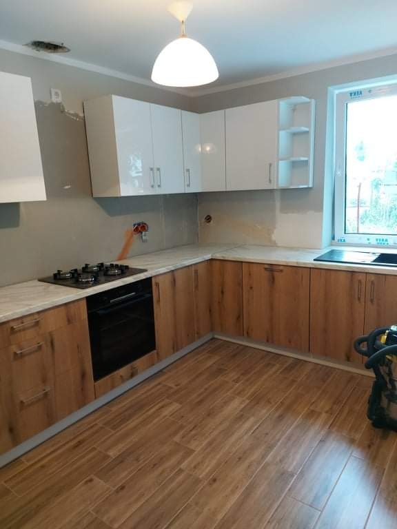 Kuchnia po remoncie: dolne szafki z drewnianym wzorem, białe, błyszczące szafki wiszące, jasny blat i podłoga z paneli imitujących drewno, widoczne niedoskonałości na ścianach przed wykończeniem.