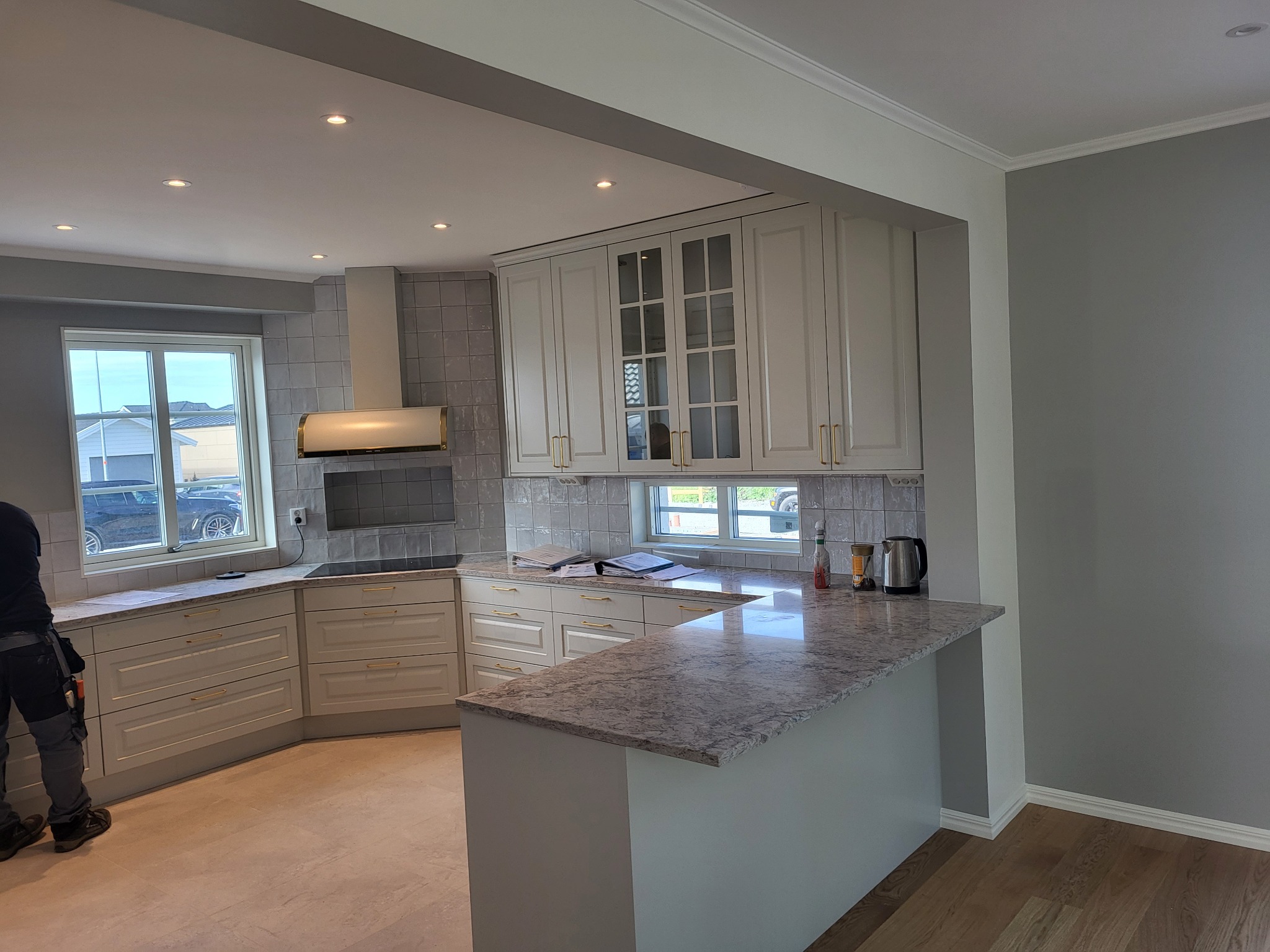 Jasna kuchnia z białymi szafkami z delikatnymi złotymi uchwytami, szarym blatem i płytkami, widok na okno i częściowo widoczna osoba w roboczych spodniach.